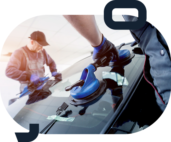 Two workers installing or repairing a car windshield, one in the foreground using a blue suction cup tool on the glass, the other in the background handling a tool with focused attention.